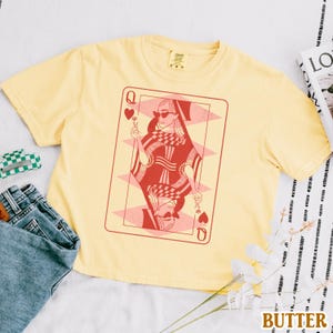 以下が含まれることがあります： ハートの女王のトランプのグラフィックがプリントされた黄色のTシャツ。女王はサングラスをかけ、赤と黒の模様のドレスを着ています。カードはピンクと赤で縁取られています。