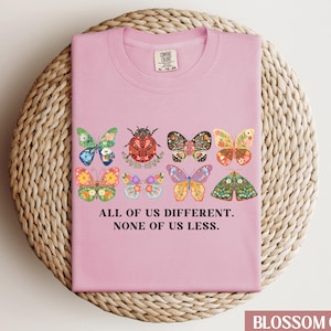 May include: Pink t-shirt with a graphic of colorful butterflies and a ladybug. The text "All of us different. None of us less." is printed on the shirt.