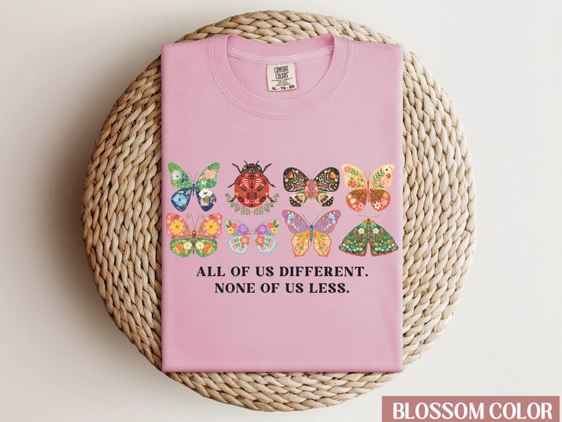 May include: Pink t-shirt with a graphic of colorful butterflies and a ladybug. The text "All of us different. None of us less." is printed on the shirt.