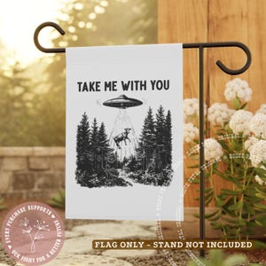 May include: White garden flag with a black and white illustration of a UFO beaming up a cow in a forest. The text "TAKE ME WITH YOU" is above the image. The flag is displayed on a black metal stand.
