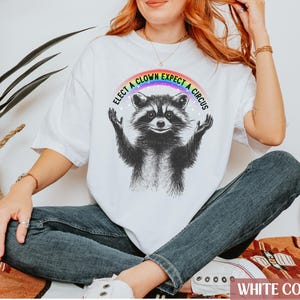 May include: White t-shirt with a rainbow and the text "Elect a Clown Expect a Circus" above a black and white illustration of a raccoon.