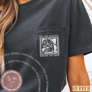 May include: Dark gray t-shirt with a pocket featuring the text "ALL YOU FASCISTS" above a floral design and "BOUND TO LOSE" below. The shirt has short sleeves and a round neckline. The brand name "PEPPER" is visible.