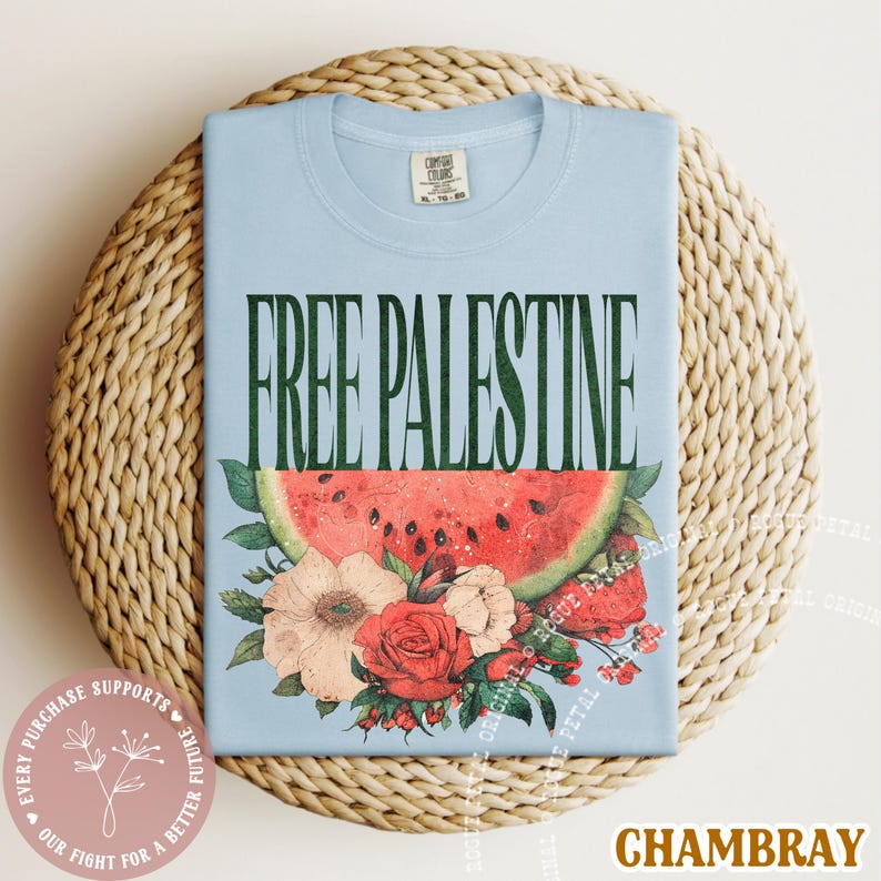May include: A light blue t-shirt with the text "FREE PALESTINE" in dark green. Below the text is a watermelon slice and floral arrangement. The shirt is displayed on a woven surface.