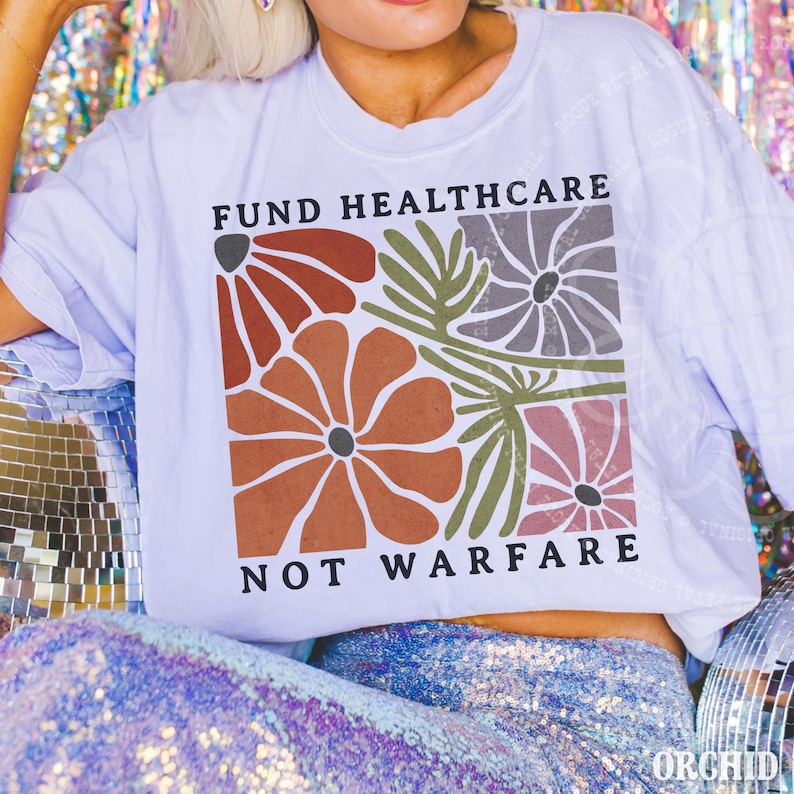 May include: A white short-sleeve t-shirt featuring a square graphic with stylized flowers in shades of rust, olive green, grey, and mauve. The text "FUND HEALTHCARE" is printed above the floral design, and "NOT WARFARE" is printed below it.