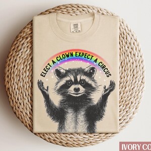 May include: A beige t-shirt with a raccoon wearing a rainbow and the text "Elect a Clown Expect a Circus".