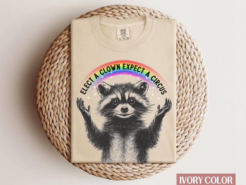 May include: A beige t-shirt with a raccoon wearing a rainbow and the text "Elect a Clown Expect a Circus".