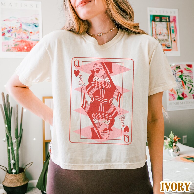 以下が含まれることがあります： ハートの女王のトランプカードの赤とピンクのグラフィックが描かれた白いTシャツ。女王はサングラスと王冠を着用しています。
