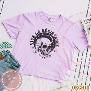 May include: Orchid-colored t-shirt with a black skull graphic, crescent moon, and stars. The text "VIVE LA R&Eacute;SISTANCE" is above the skull. The shirt is folded with a pair of jeans and a hair clip.