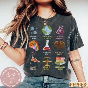 May include: Dark gray t-shirt with various graphics and text, including "Vaccines Work," "There is no Planet B," and "Black Lives Matter." Other graphics include a globe, a rainbow, and a beaker. The shirt also has the text "Love is Love" and "Equality for All."