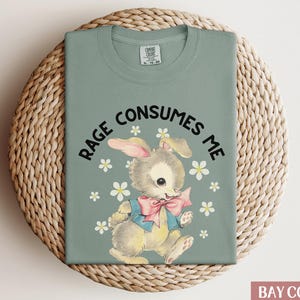 Op de afbeelding: Een saliegroen T-shirt met de tekst "Rage Consumes Me" in zwart gedrukt. Een cartoonkonijn met een roze strik is gecentreerd op het shirt, omringd door witte bloemen.