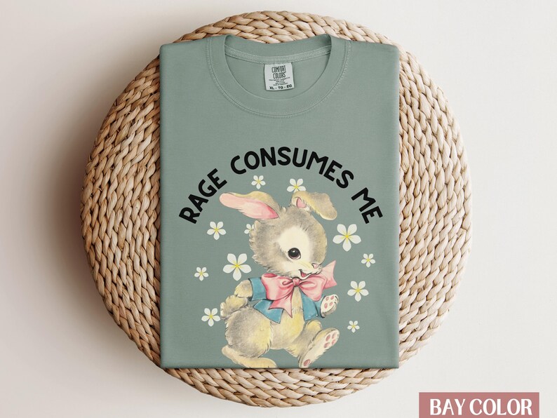 Op de afbeelding: Een saliegroen T-shirt met de tekst "Rage Consumes Me" in zwart gedrukt. Een cartoonkonijn met een roze strik is gecentreerd op het shirt, omringd door witte bloemen.