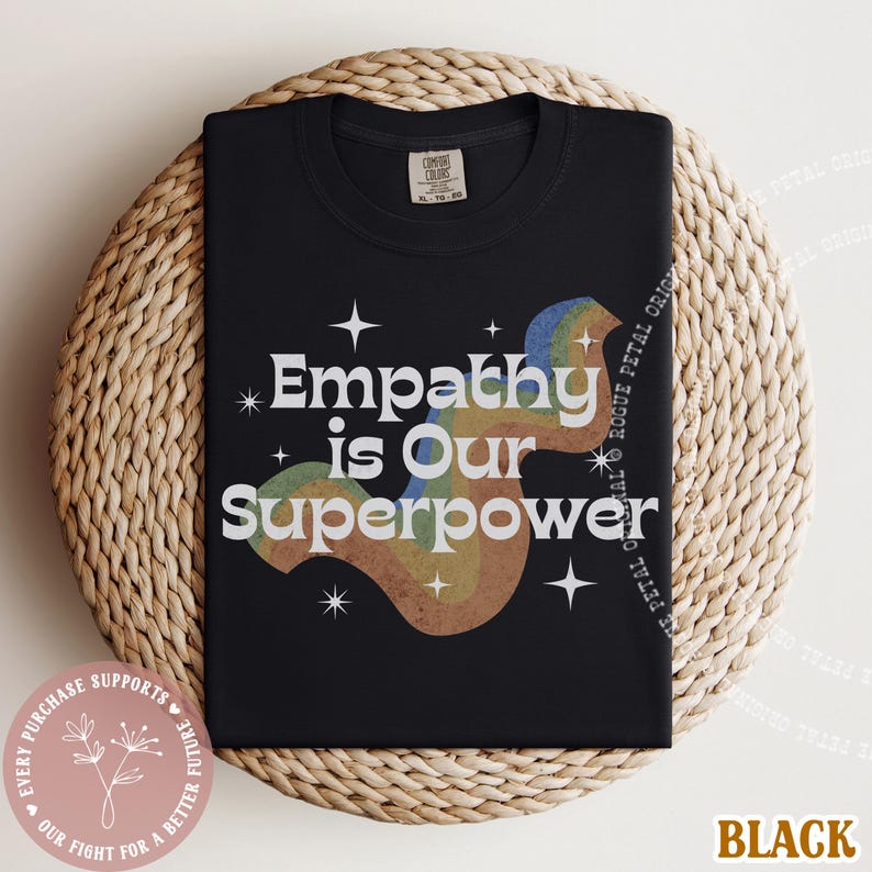 Puede incluir: Camiseta negra con la frase blanca "Empathy is Our Superpower", acompa&ntilde;ada de un dise&ntilde;o de arco&iacute;ris. La camiseta est&aacute; doblada sobre una superficie tejida. La palabra "Black" est&aacute; en la parte inferior derecha.
