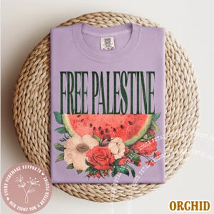 May include: Orchid-colored t-shirt with the text "FREE PALESTINE" in dark green. A watermelon slice and floral arrangement are below the text. The shirt is folded on a woven surface.