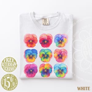 May include: A white t-shirt featuring a grid of nine watercolor-style pansy flowers in various vibrant colors including orange, red, blue, yellow, and purple. The shirt has a Comfort Colors brand label at the inner neckline and is size M.