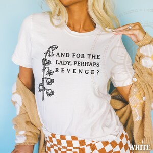 以下が含まれることがあります： 左側にスズランの茎の黒いグラフィックが描かれた白い半袖Tシャツ。花のイラストの右側には「AND FOR THE LADY, PERHAPS REVENGE?」というテキストが記されています。Tシャツはベージュのカーディガンと合わせて着用されています。