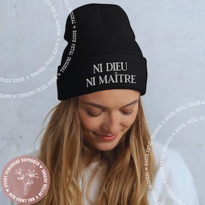 May include: Black beanie with the French text "NI DIEU NI MAÎTRE" embroidered in white. The beanie is made of a soft, knitted material. The image also includes a circular graphic with text that reads "EVERY PURCHASE SUPPORTS OUR FIGHT FOR A BETTER FUTURE."