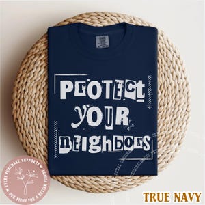 May include: True navy blue t-shirt with the text "PROTECT YOUR NEIGHBORS" in white block letters. The shirt is folded on a woven, circular mat. The phrase "EVERY PURCHASE SUPPORTS OUR FIGHT FOR A BETTER FUTURE" is printed on a pink circle.