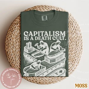 K&ouml;nnte beinhalten: Ein moosgr&uuml;nes T-Shirt mit dem wei&szlig;en Text "CAPITALISM IS A DEATH CULT". Die Grafik zeigt Arbeiter, die S&auml;rge bauen. Das Shirt ist auf einer gewebten Matte gefaltet. Ein rosa Aufkleber mit Text befindet sich unten links.