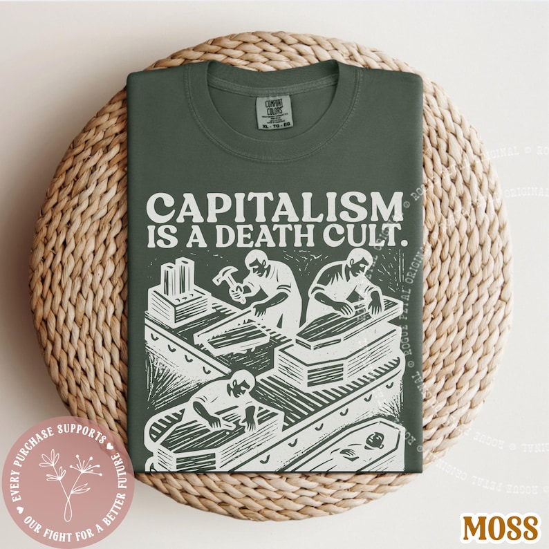 K&ouml;nnte beinhalten: Ein moosgr&uuml;nes T-Shirt mit dem wei&szlig;en Text "CAPITALISM IS A DEATH CULT". Die Grafik zeigt Arbeiter, die S&auml;rge bauen. Das Shirt ist auf einer gewebten Matte gefaltet. Ein rosa Aufkleber mit Text befindet sich unten links.
