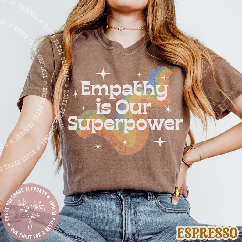 Puede incluir: Camiseta marr&oacute;n con la frase blanca "Empathy is Our Superpower" y un dise&ntilde;o de arco&iacute;ris. La camiseta tiene cuello en V y mangas cortas. El dise&ntilde;o incluye estrellas.