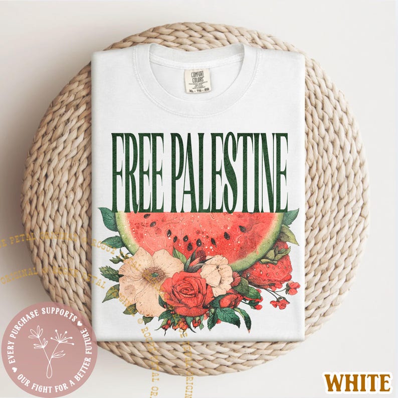 May include: White t-shirt with the text "FREE PALESTINE" in green, above a watermelon slice and floral arrangement. The design includes red roses, peach flowers, and green leaves. The shirt is folded on a woven surface.