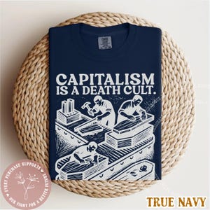 K&ouml;nnte beinhalten: Marineblaues T-Shirt mit dem Text "CAPITALISM IS A DEATH CULT." und einer wei&szlig;en Grafik von Personen, die S&auml;rge bauen. Das T-Shirt liegt auf einer geflochtenen Oberfl&auml;che.