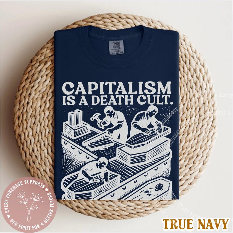 K&ouml;nnte beinhalten: Marineblaues T-Shirt mit dem Text "CAPITALISM IS A DEATH CULT." und einer wei&szlig;en Grafik von Personen, die S&auml;rge bauen. Das T-Shirt liegt auf einer geflochtenen Oberfl&auml;che.