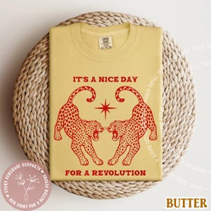 May include: Butter-colored t-shirt with a red graphic of two leopards facing each other, a star, and the text "IT'S A NICE DAY FOR A REVOLUTION." The shirt is displayed on a woven surface.