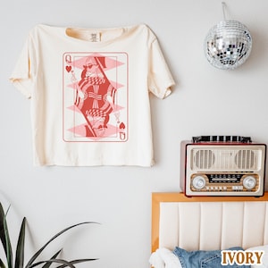 以下が含まれることがあります： 赤いスペードの女王のグラフィックがプリントされた白いクロップドTシャツ。グラフィックには、サングラスと王冠をつけた女性が描かれています。