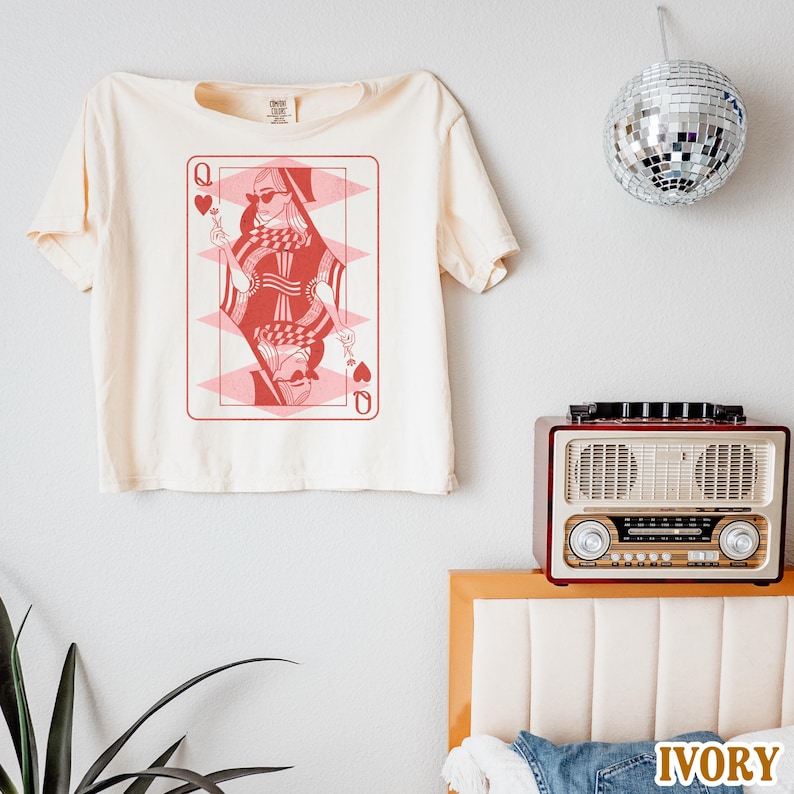 以下が含まれることがあります： 赤いスペードの女王のグラフィックがプリントされた白いクロップドTシャツ。グラフィックには、サングラスと王冠をつけた女性が描かれています。