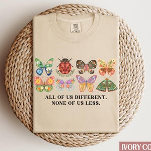 May include: A beige t-shirt with a graphic of colorful butterflies and a ladybug. The text "All of us different. None of us less." is printed below the graphic.