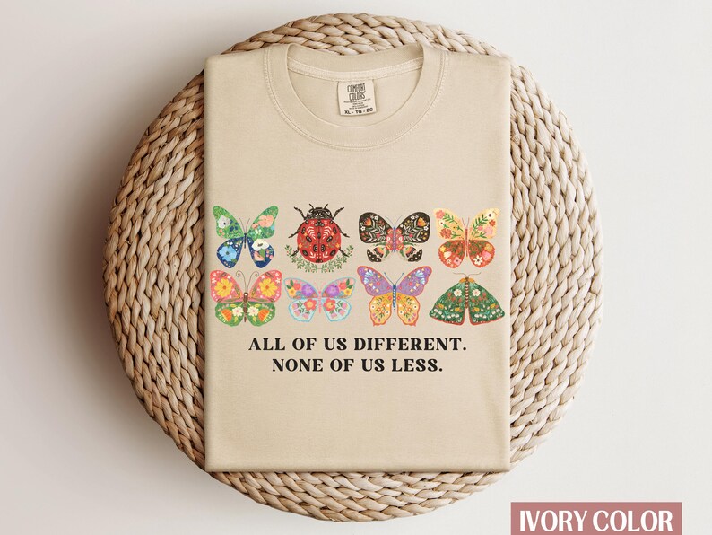 May include: A beige t-shirt with a graphic of colorful butterflies and a ladybug. The text "All of us different. None of us less." is printed below the graphic.