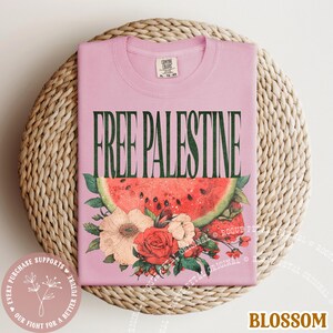 May include: A light pink t-shirt with the text "FREE PALESTINE" in green. Below the text is a watermelon slice and floral arrangement. The shirt is folded on a woven surface.