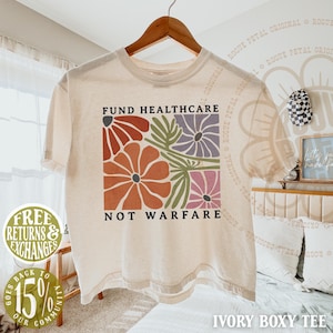 May include: An ivory boxy t-shirt featuring a graphic print of stylized flowers in shades of red, orange, purple, and pink with green leaves. The text "FUND HEALTHCARE" is printed above the floral design and "NOT WARFARE" is printed below it.