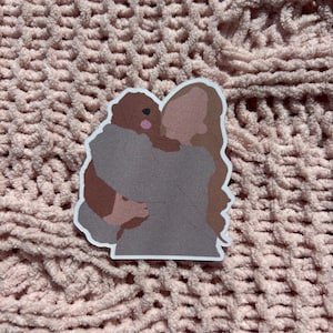 Może przedstawiać: Naklejka przedstawiająca osobę obejmującą brązowego psa. Osoba ma na sobie szarą koszulę, a pies jest w jej ramionach. Naklejka ma białą obwódkę i jest umieszczona na różowym, teksturowanym tle.