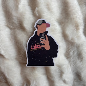 May include: A sticker depicting a person in a black hoodie and a baseball cap, taking a selfie. The hoodie has pink and white text and small yellow stars. The cap is grey and pink.
