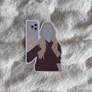 May include: A sticker featuring a person taking a selfie with a phone. The person has long blonde hair and a blue face mask. The phone is gray and pink, and the person is wearing a dark brown top.