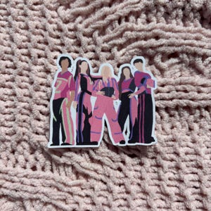 May include: A sticker depicting a group of six people in stylized outfits. The outfits are mainly pink, black, and white, with striped and patterned details. The sticker is on a pink, textured surface.