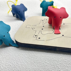 Peut inclure: Quatre porte-clés en cuir en forme d'éléphant dans différentes couleurs : bleu, rouge et deux nuances de bleu. Les porte-clés sont sur une planche à découper en bois avec une découpe en forme d'éléphant.