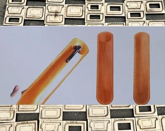 May include: A collection of wooden die-cut templates for leather pen cases, alongside finished leather pen holders in a warm tan color. One case holds a pen. The templates are arranged in a grid pattern.