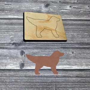 Puede incluir: Un troquel de madera de un perro con un contorno negro y el número 473. Un recorte de perro de cuero marrón está debajo. El fondo es una superficie de madera gris.