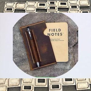 Könnte beinhalten: Eine braune Leder-Notizbuchhülle mit Stifthalter, die ein Field Notes-Notizbuch enthält. Die Hülle hat ein Used-Finish und weiße Nähte. Ein schwarzer Stift befindet sich im Halter. Das Notizbuch ist beige mit schwarzem Text.