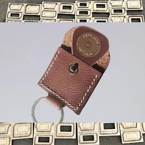 Può includere: Un porta plettri in pelle marrone con portachiavi. Il supporto presenta cuciture bianche e una chiusura a scatto. Un plettro per chitarra con un design circolare è visibile all'interno. Lo sfondo include ritagli di legno.