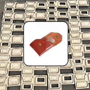 May include: A small, reddish-brown leather coin pouch with a brass snap closure, containing two metallic coins. The pouch is displayed against a background of numerous rectangular wooden cutting dies used for leather crafting projects.