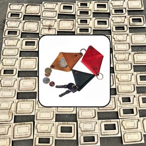 May include: Three triangular leather coin pouches in tan, red, and black, each featuring a metal snap closure and a key ring. The tan pouch holds a silver coin, while the black pouch is attached to a set of metal keys. Several loose coins lie nearby.