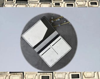 May include: A handmade white and black leather wallet with a gray interior, open to display card slots and a pocket. The wallet is on a gray surface, surrounded by wooden die-cut shapes.