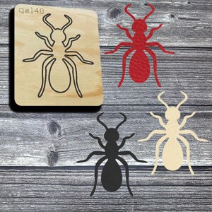 May include: A wooden die cut with an ant outline and the text "qw140". The image also shows three ant cutouts in red, black, and cream colors. The cutouts are made of leather or a similar material. The background is a gray wood grain.