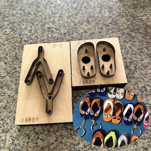 Puede incluir: Dos troqueles de corte de madera para la elaboración de sandalias de cuero. Los troqueles están etiquetados como "2-1" y "2-2". El troquel etiquetado como "2-1" tiene dos formas de sandalias. El troquel etiquetado como "2-2" tiene una forma más compleja. La imagen también muestra un primer plano de varias sandalias de cuero que se han cortado utilizando los troqueles.