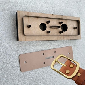 Puede incluir: Un cinturón de cuero marrón con una hebilla de metal dorado. La hebilla es rectangular con esquinas redondeadas y una superficie lisa. El cinturón se muestra junto a una herramienta de madera con varios agujeros, probablemente utilizada para la elaboración de artículos de cuero.