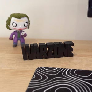 Pode incluir: Letras de plástico preto soletrando a palavra "WARZONE" em uma superfície de madeira.